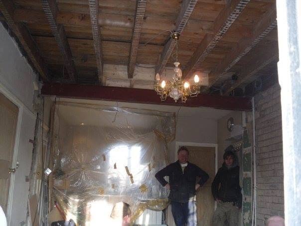 A room stripped back to the joists with a lit chandelier hanging, two workers in the doorway
